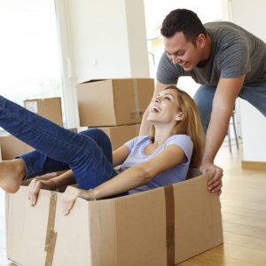 A young, cheerful, caucasian couple is having fun and playing in their new home. The young woman is smiling and sitting in a box while her partner is pushing the box. In the backround we can see some boxes and the house interior.
